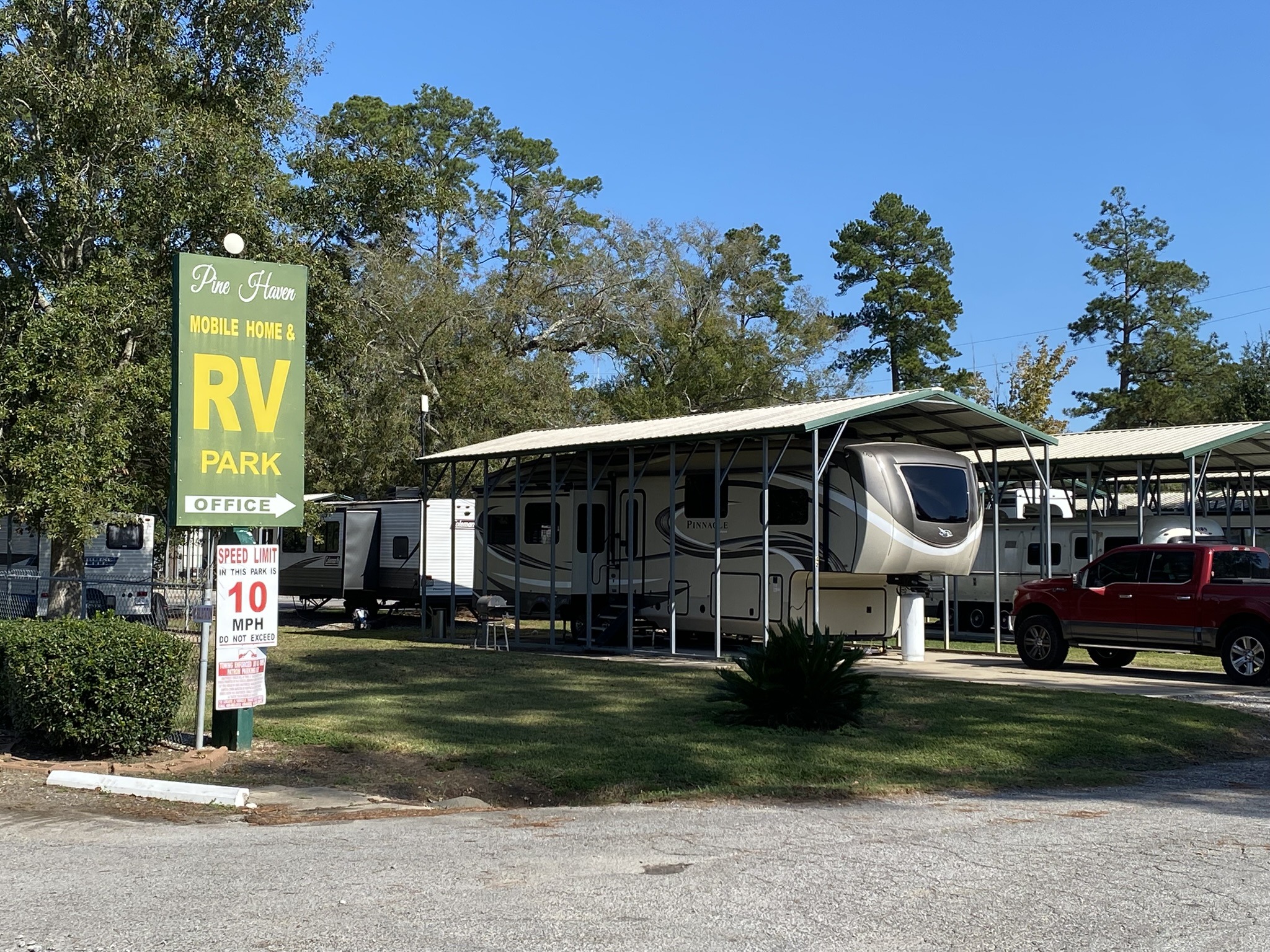 RV and mobile home park under clear skies. A sign reads "Pine Haven Mobile Home & RV Park." Covered RVs and a red truck are visible. Trees in the background.