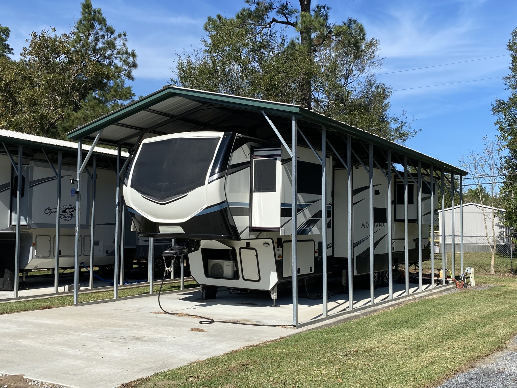 A fifth-wheel RV is parked under a metal carport on a sunny day. Tall green trees and blue sky are in the background. The setting is calm and tidy.