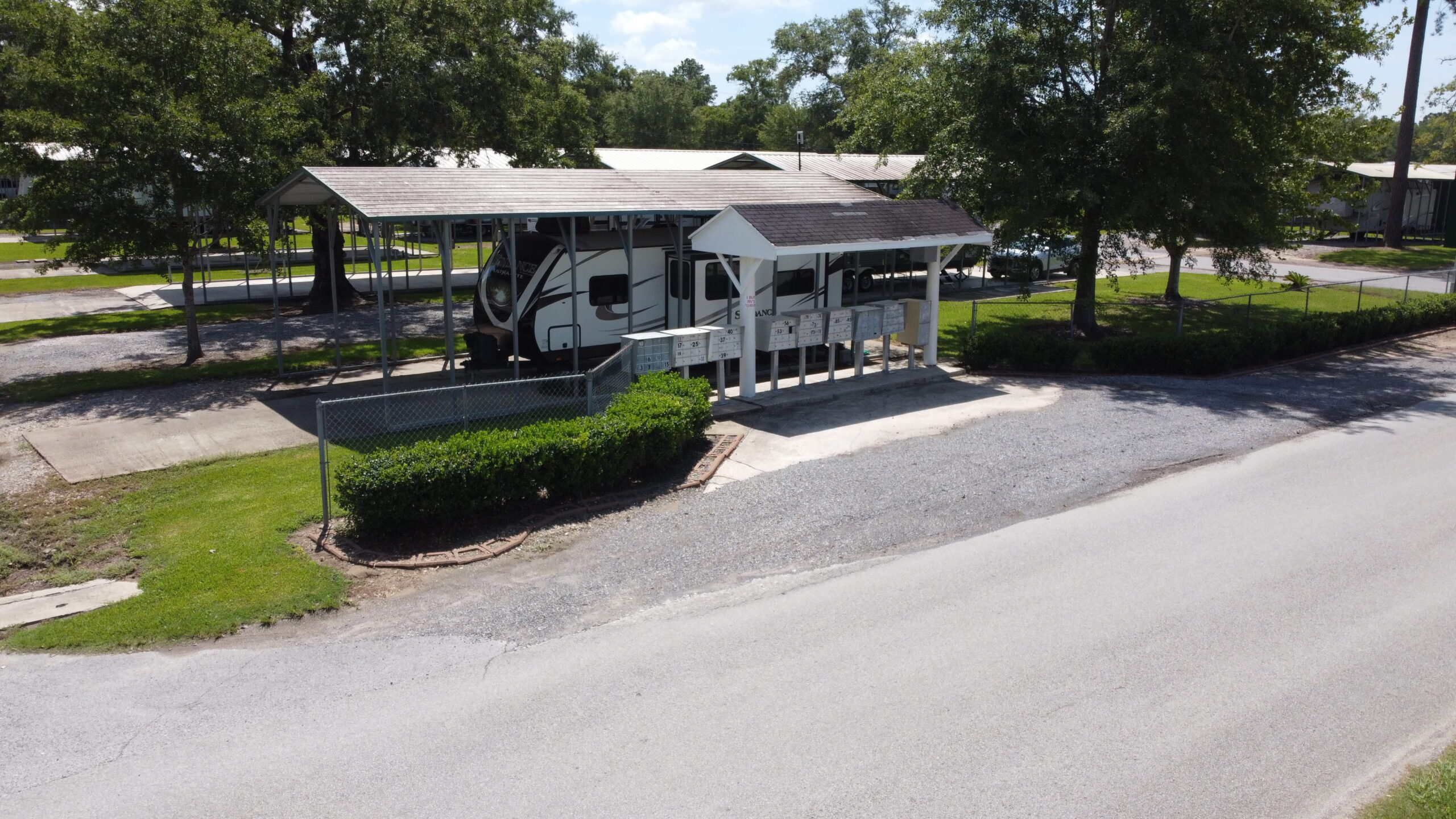 A covered RV parked in a driveway next to a road, surrounded by green trees and shrubs. The area is peaceful, with a clear, sunny sky.